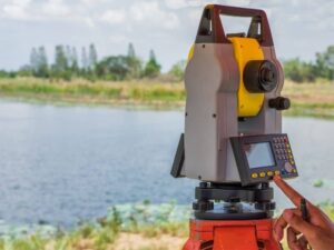 Total station surveying instrument on a tripod beside a body of water, with a hand on its control panel.